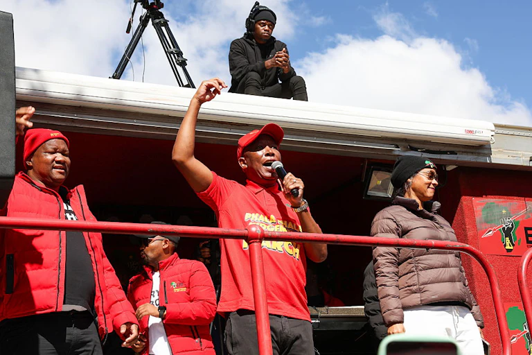 Man in red shirt and cap with arm raised, speaking into a microphone