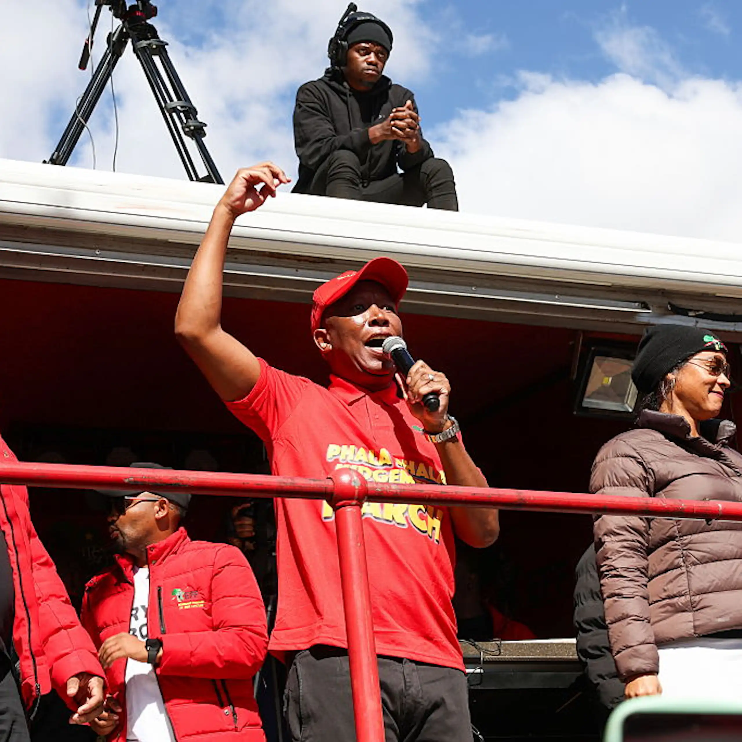Man in red shirt and cap with arm raised, speaking into a microphone