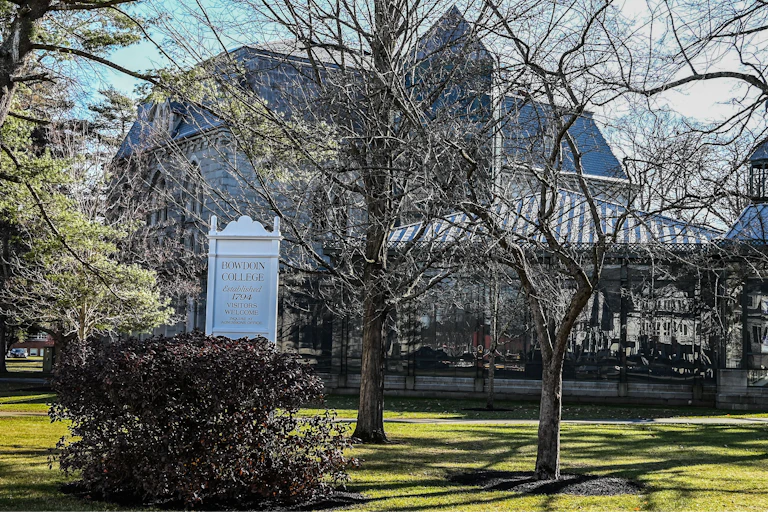 A white sign says 'Bowdoin College' in front of a building and trees on a grassy field.