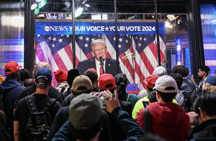 Several people wearing baseball hats watch a man speak on TV.