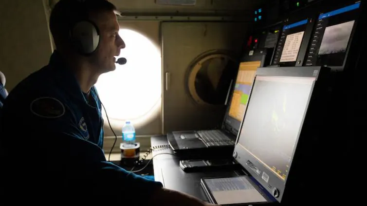Los pronósticos de huracanes mejoran dramáticamente, salvando vidas, pero los recortes federales amenazan con llevar a la NOAA al límite 1 Un científico con traje de vuelo se sienta frente a una computadora en un avión y habla a través de unos auriculares.