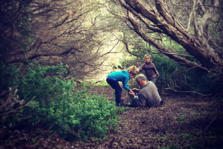 A man lies on the ground in the bush talking to two children.