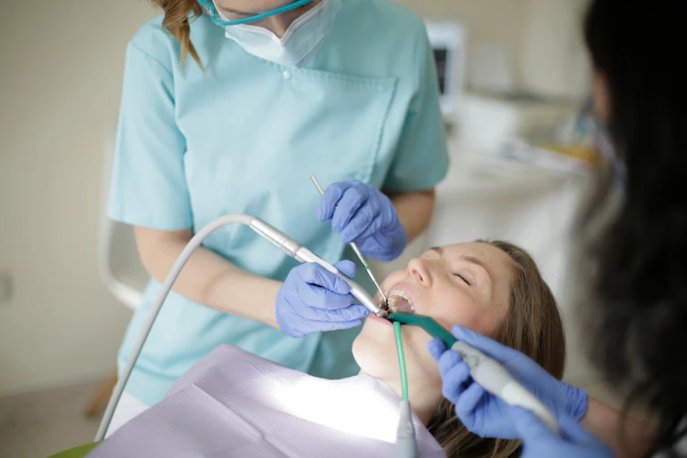 Female dentist working on mouth of female patient, lying down in chair, dental assistant helping