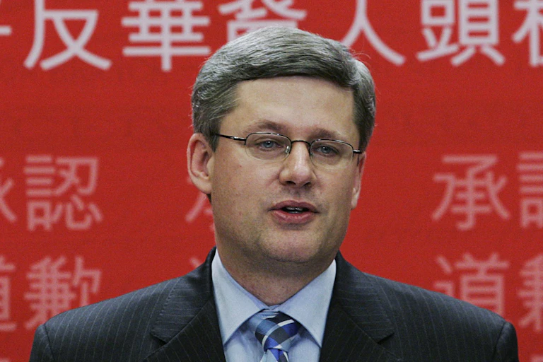 A man with dark grey hair wearing glasses stands in front of a red banner with Chinese characters.