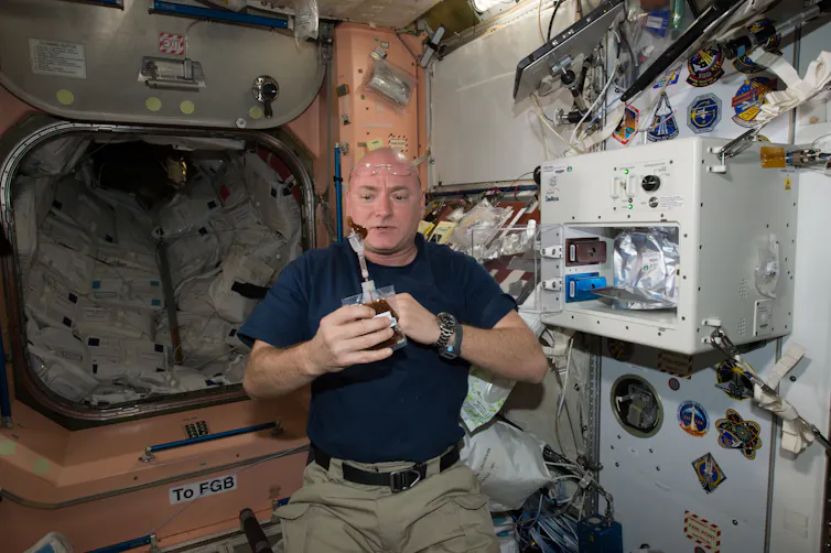 An astronaut holding a plastic pouch with an attached straw. A brown bead of liquid is floating in the air outside the straw.