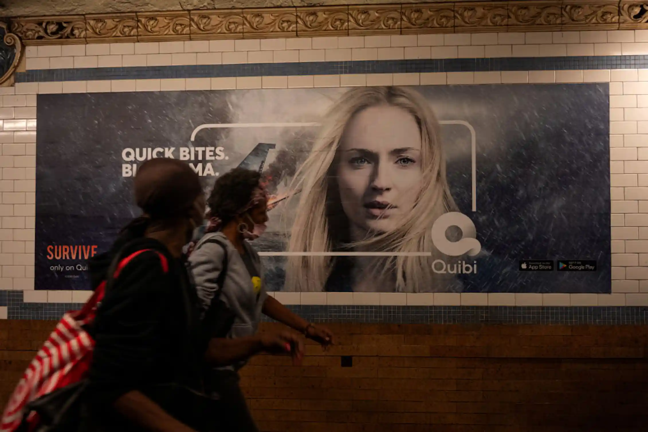 People wearing masks in a subway station walk past an advertisement featuring a young woman and the text 'quick bites, big drama.'