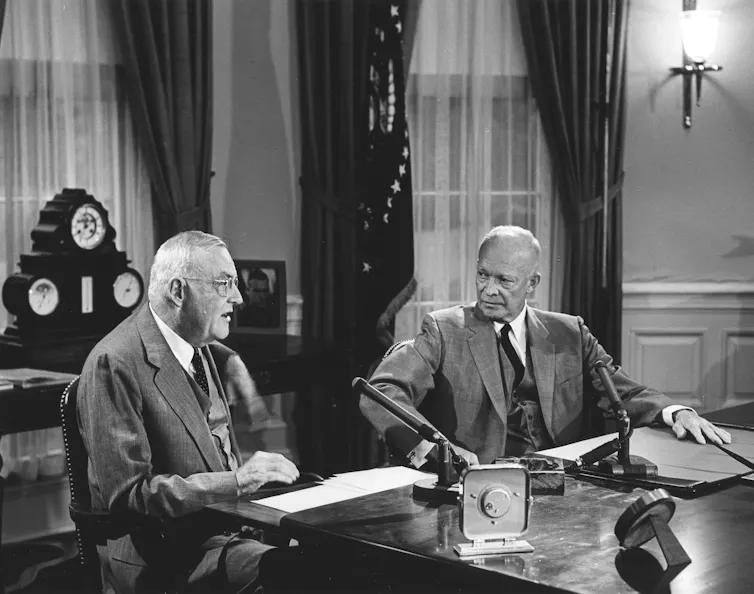 Two men in suits and ties talk while seated in front of a table.