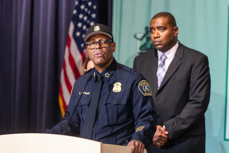 A Black man wearing a police uniform stands behind podium.