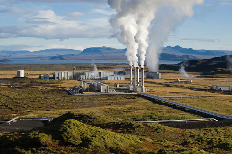 Steam rising from a geothermal plant, pipes and industrial facility with sea behind.