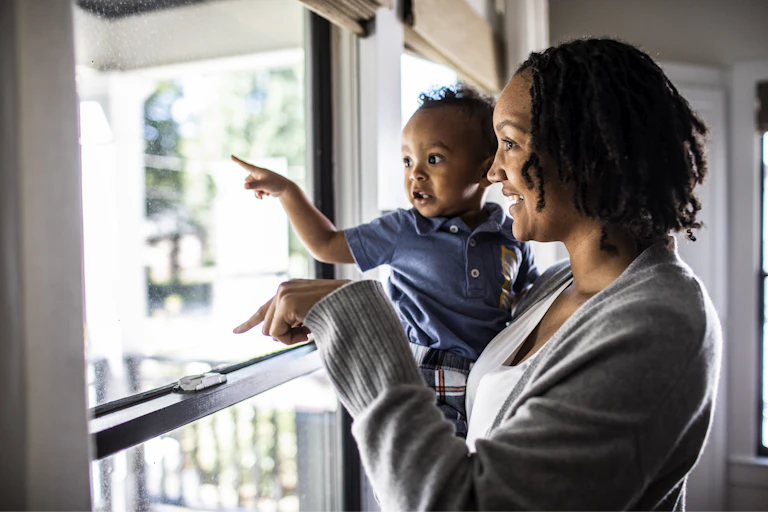 A woman holds a small child and they both point, looking out the window together.