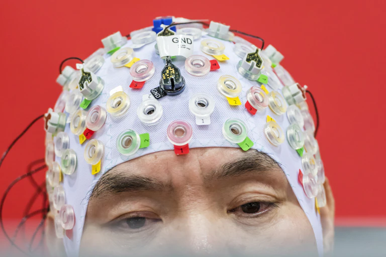 Man wearing a cap-type device with lots of colourful electrodes and wires looks at a computer screen.