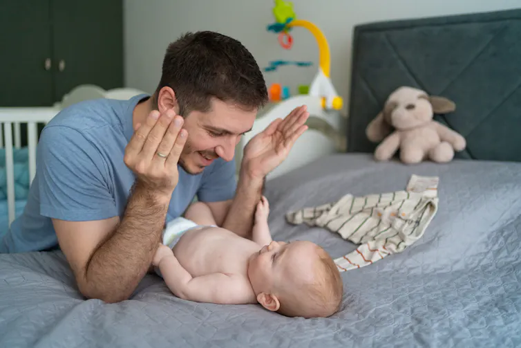 A man holds his hands away from his face and leans over a small baby lying on a bed and smiles.