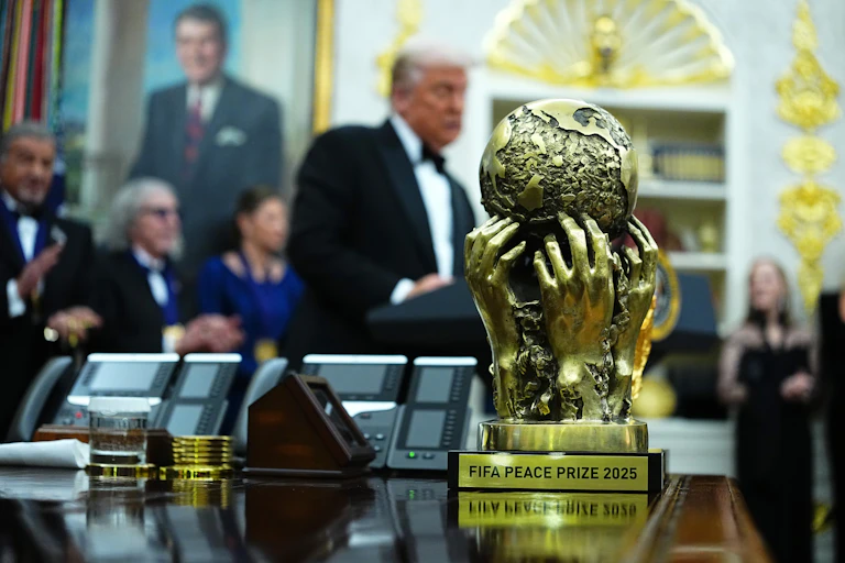 A shiny gold trophy of a globe held up by human hands, in the background a blurry shot of men in suits, one with white hair standing and talking.