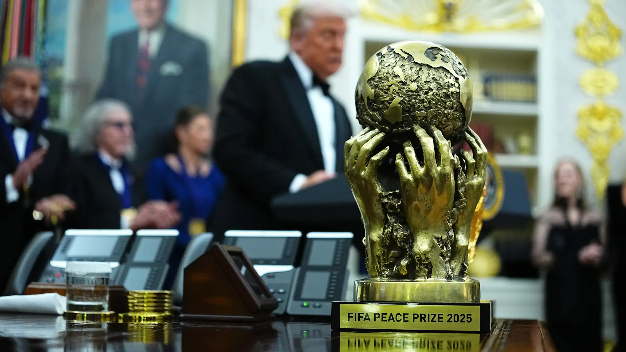 A shiny gold trophy of a globe held up by human hands, in the background a blurry shot of men in suits, one with white hair standing and talking.