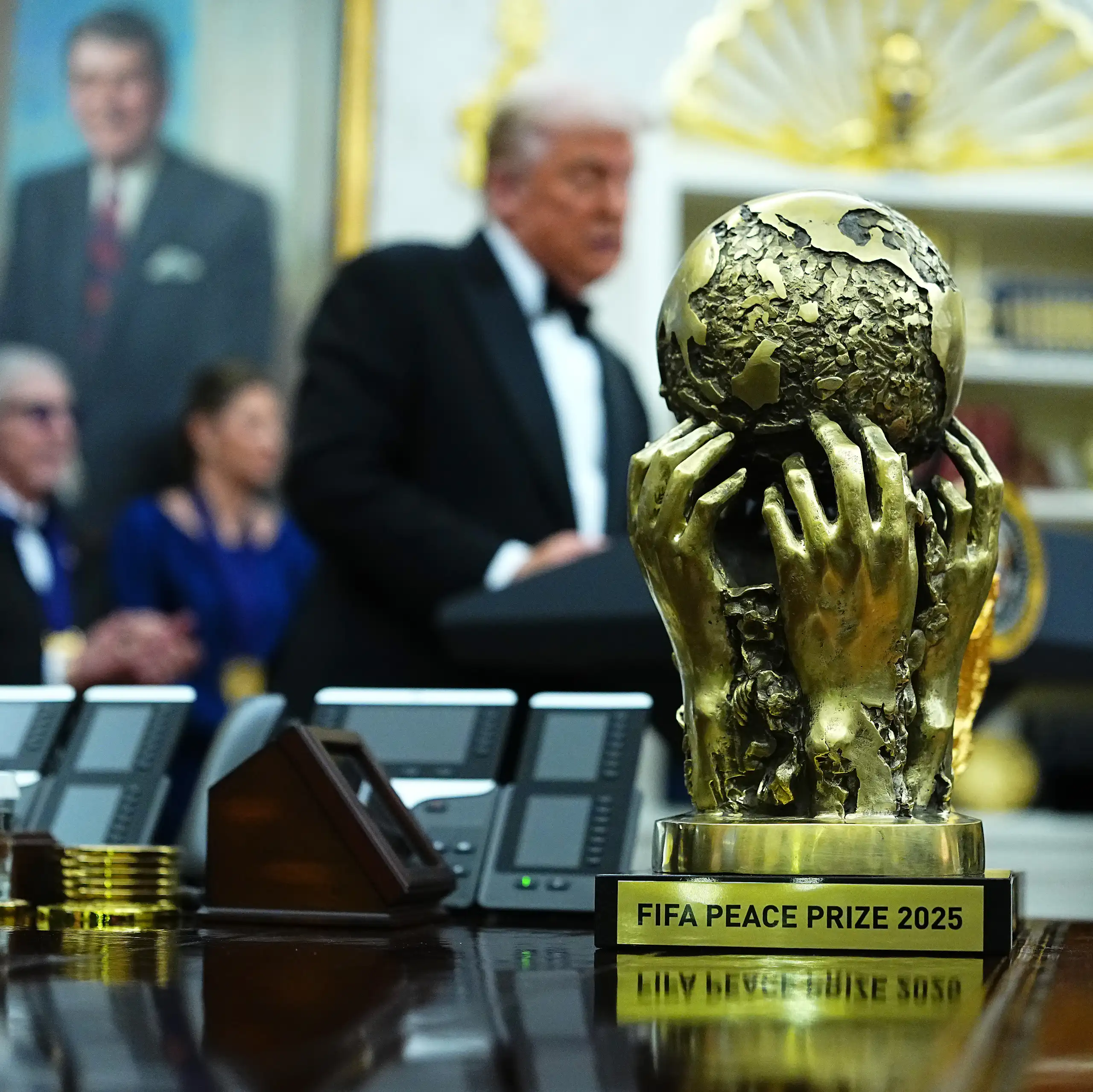 A shiny gold trophy of a globe held up by human hands, in the background a blurry shot of men in suits, one with white hair standing and talking.