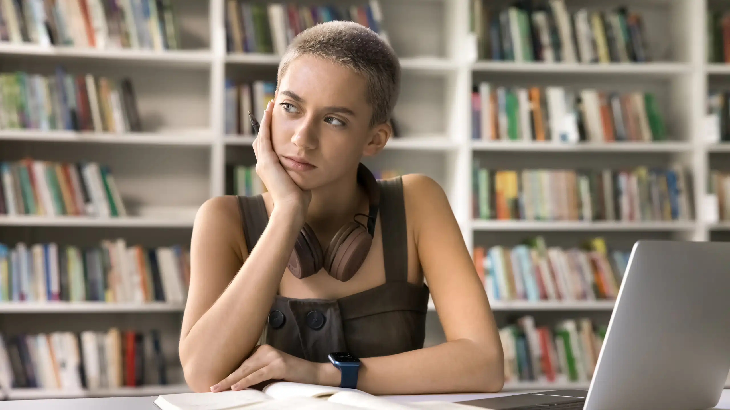Joven con aspecto pensativo sentada con un libro abieto y un ordenador