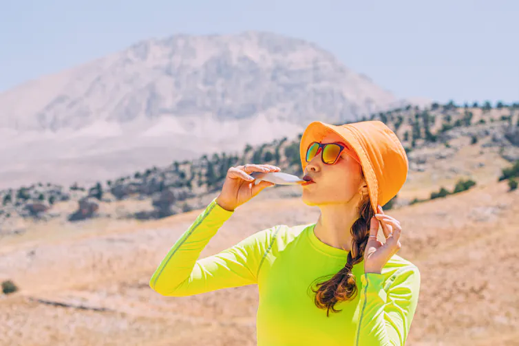 A woman wearing a hat and athletic gear slurps down an energy gel on a sunny day in the desert.