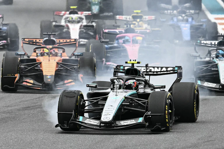 Several open-wheel race cars jostle for position, emitting a cloud including smoke from tires and exhaust from their engines.