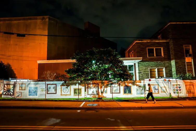 A tree stands in front of a fence covered with lit-up signs, standing in front of a few buildings at night.