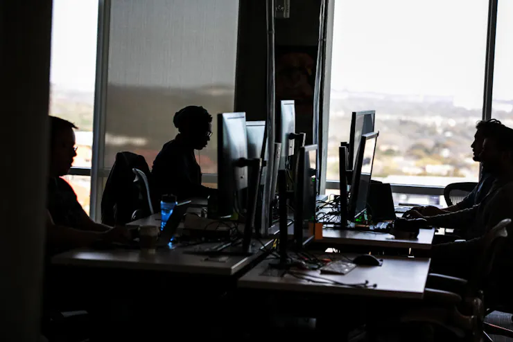 A dark photograph shows a handful of people sitting at large computer screens in a room with a large windows.