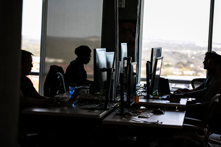 A dark photograph shows a handful of people sitting at large computer screens in a room with a large windows.
