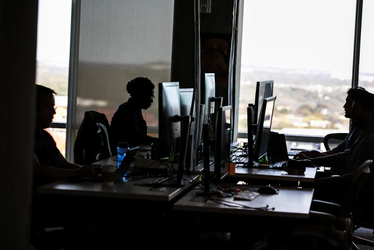 A dark photograph shows a handful of people sitting at large computer screens in a room with a large windows.