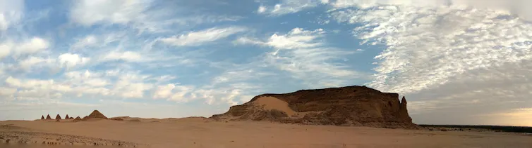 Panoramic view of a sandy landscape with a large mesa on the right and smaller pyramids in the distance, all against a blue sky.