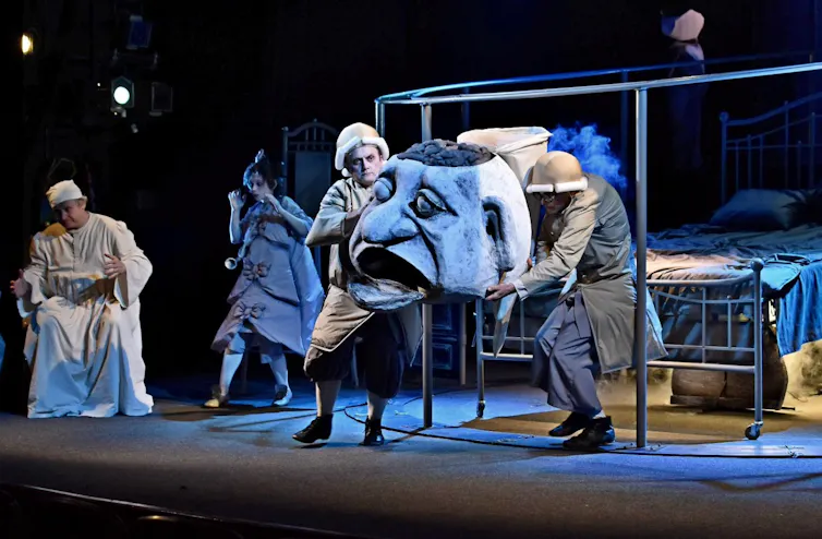 A theatre troupe on stage with the large head of a puppet.