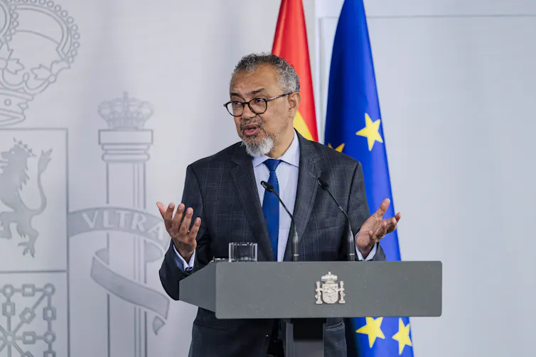 A man in a grey suit speaking at a podium with flags behind him