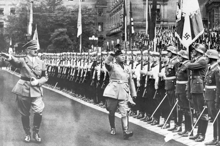 Hitler and Mussolini walk past a row of saluting soldiers, returning the salute