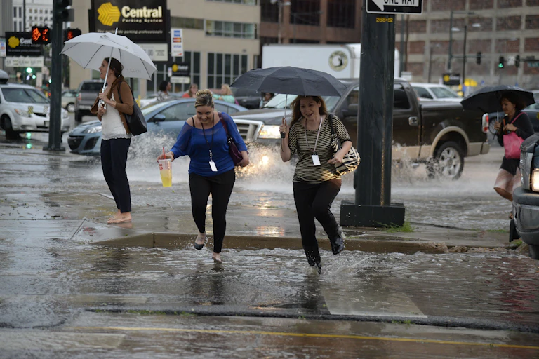 Two women carrying umbrellas laugh as they run through ankle-deep puddles in a city street.