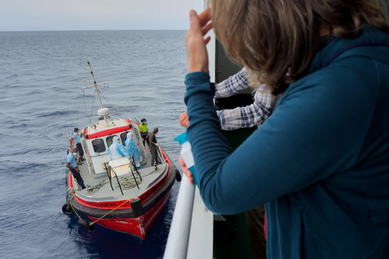 Shoulders of passengers on deck of ship, a small boat docking below with people dressed in personal protective gear