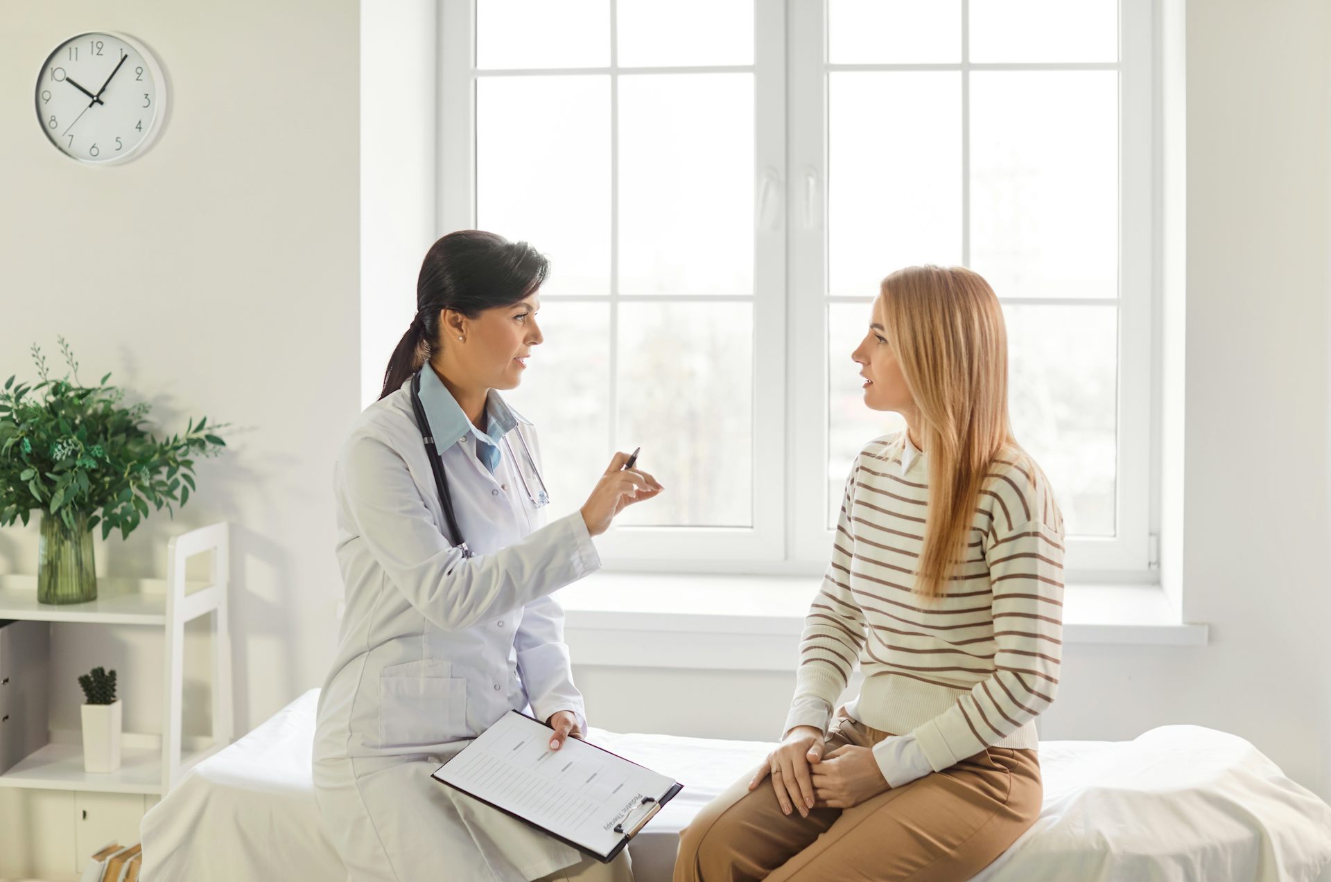 A female doctor speaks with her young, female patient who is sitting on an examination bed.