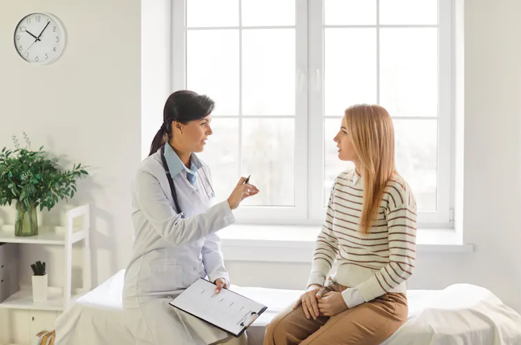 A female doctor speaks with her young, female patient who is sitting on an examination bed.
