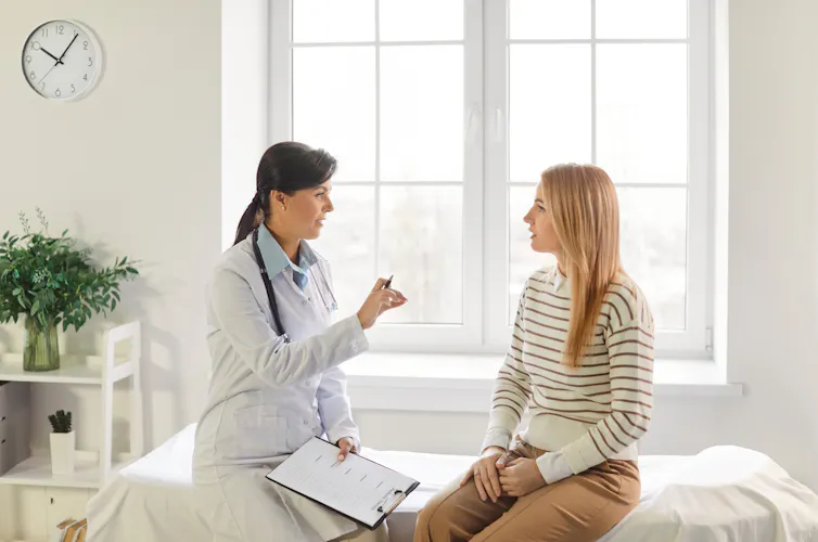 A female doctor speaks with her young, female patient who is sitting on an examination bed.