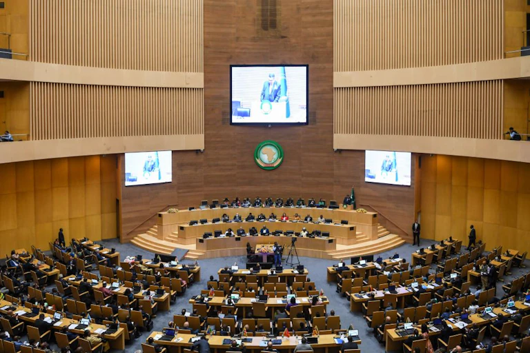 People seated in a large auditorium, with screens on the wall and a logo of the map of Africa
