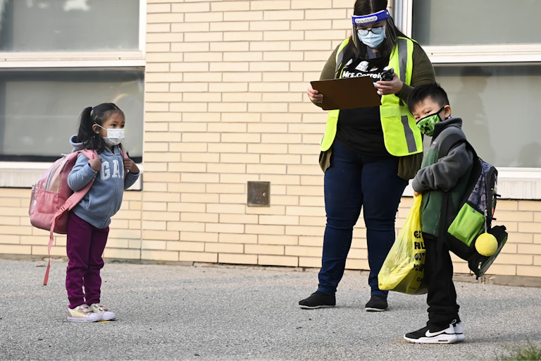 Two children seen in face masks wearing backpacks next to an adult in a safety vest and mask holding a clip board.