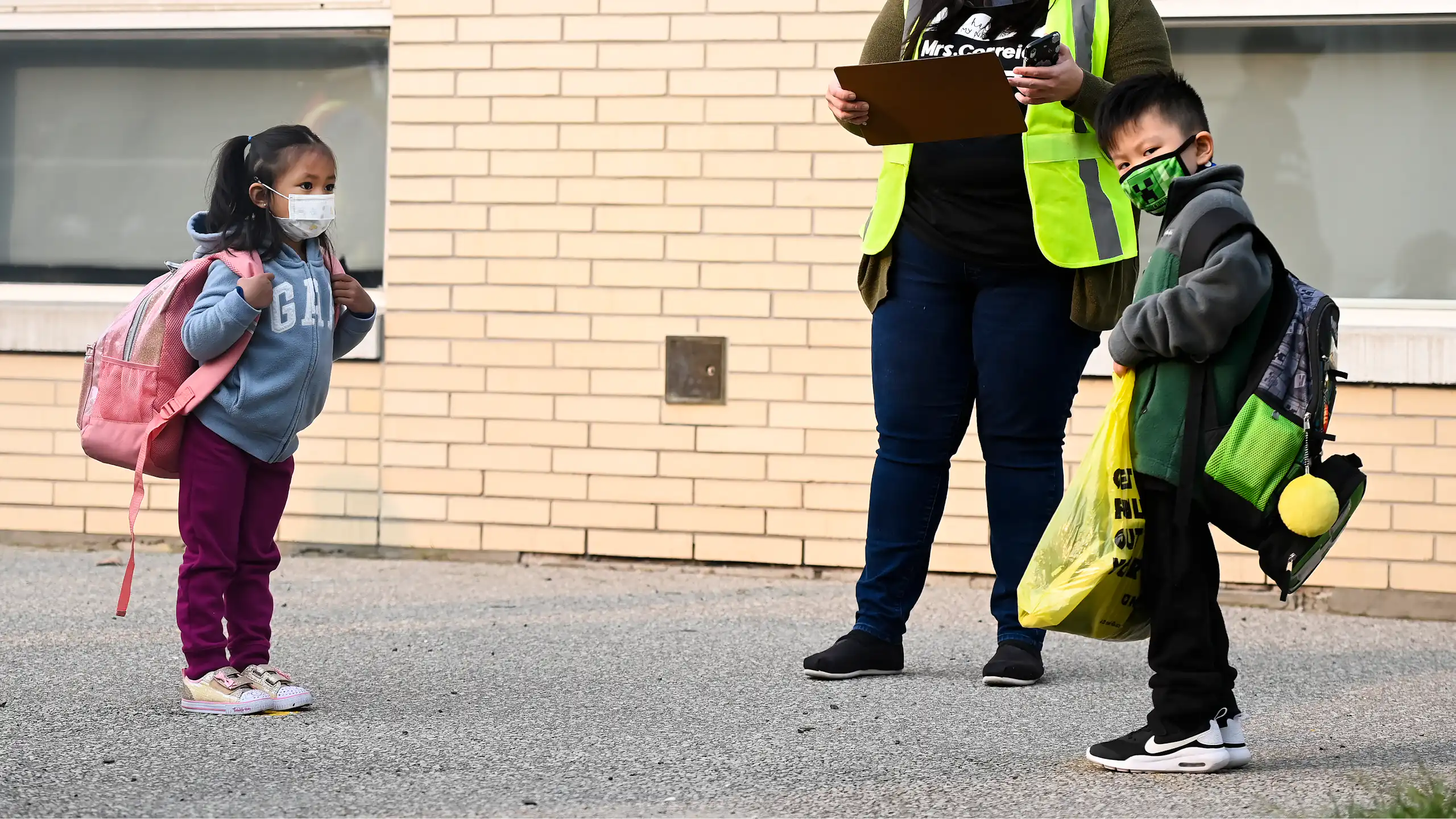Two children seen in face masks wearing backpacks next to an adult in a safety vest and mask holding a clip board.