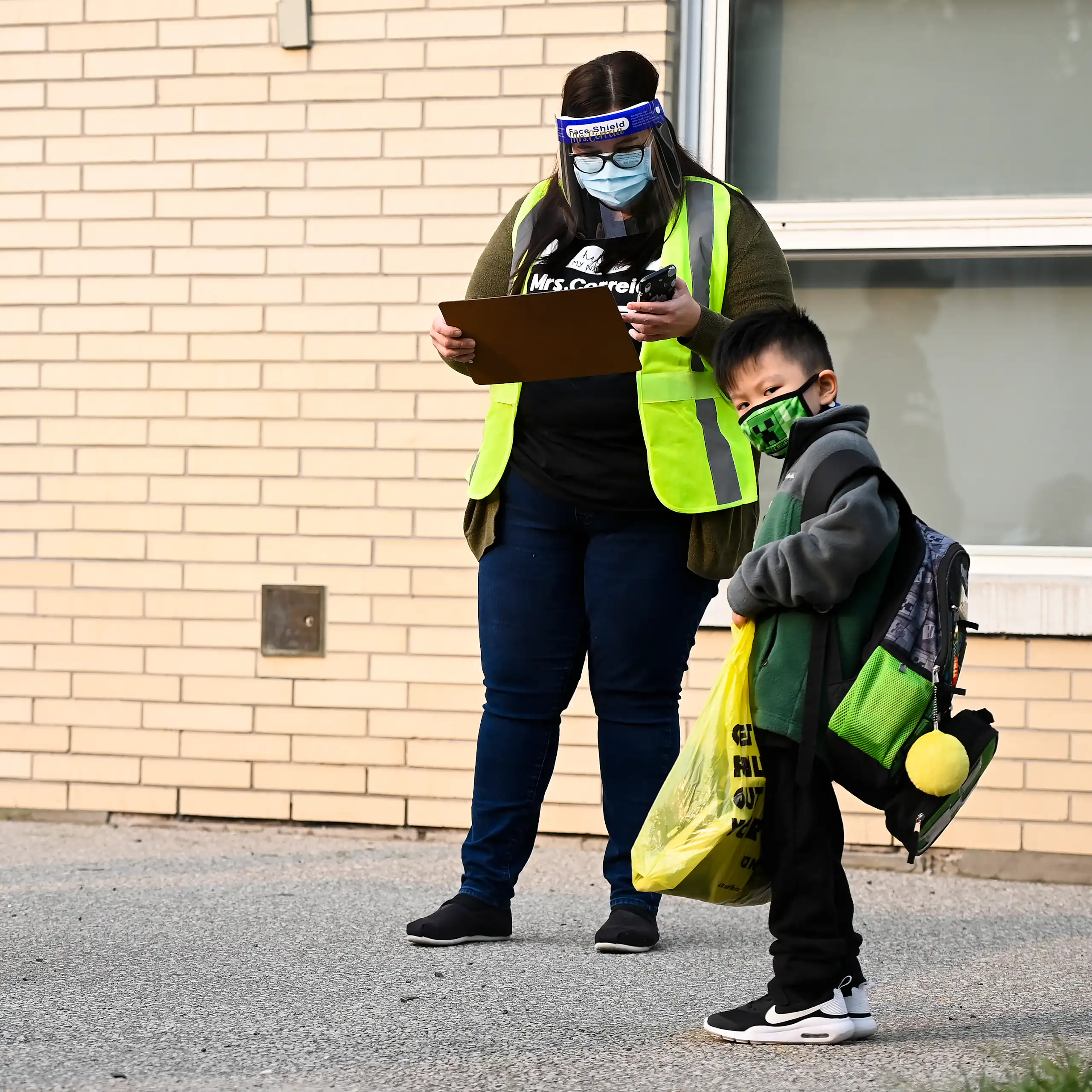 Two children seen in face masks wearing backpacks next to an adult in a safety vest and mask holding a clip board.