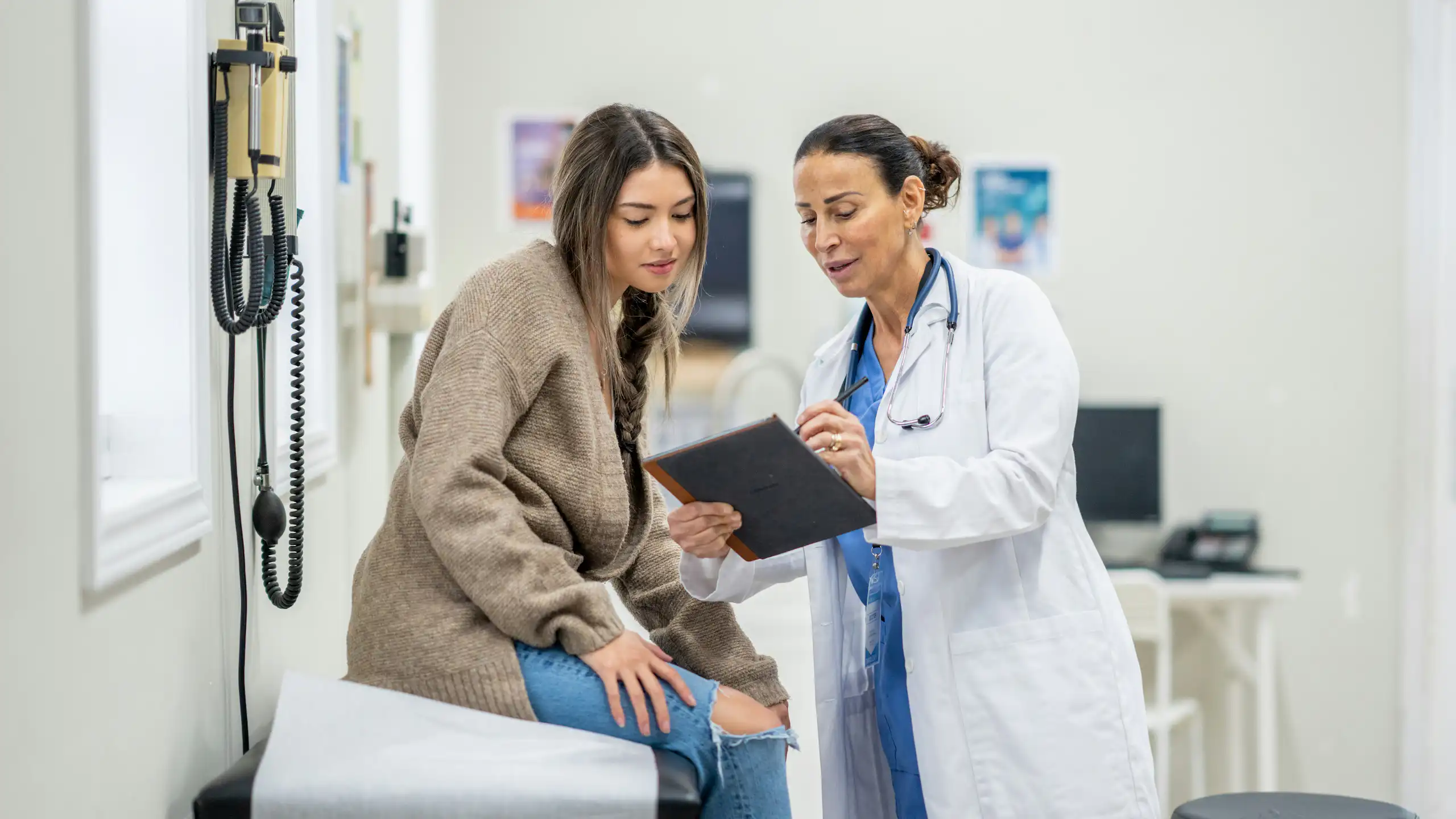 A woman in a white coat with a stethoscope speaking with a young woman in a clinic exam room