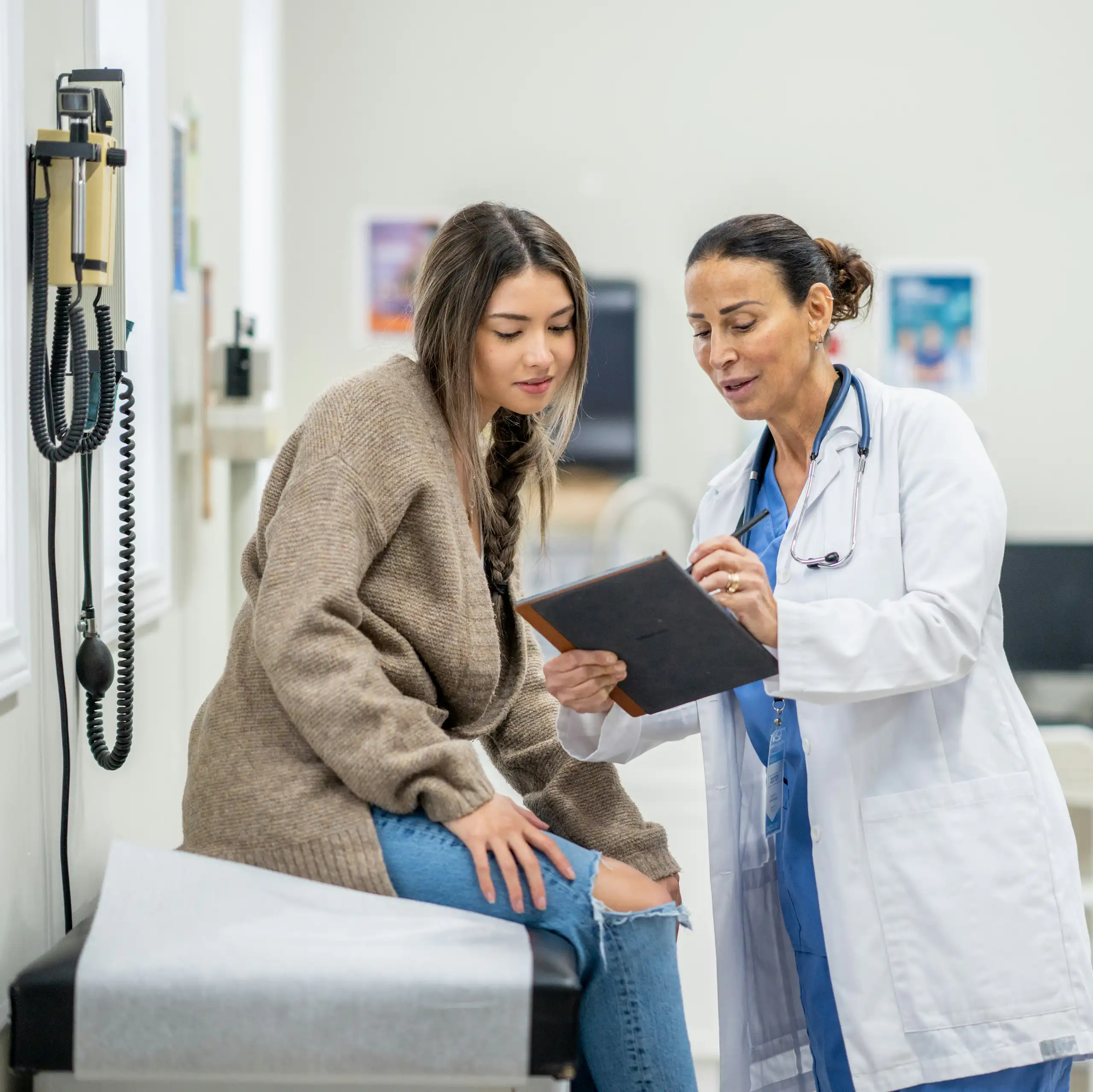 A woman in a white coat with a stethoscope speaking with a young woman in a clinic exam room