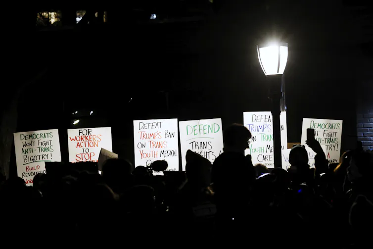 Protestors holding signs next to a street lamp at night, two of which read 'DEFEAT TRUMP'S BAN ON TRANS YOUTH MEDICAL CARE'