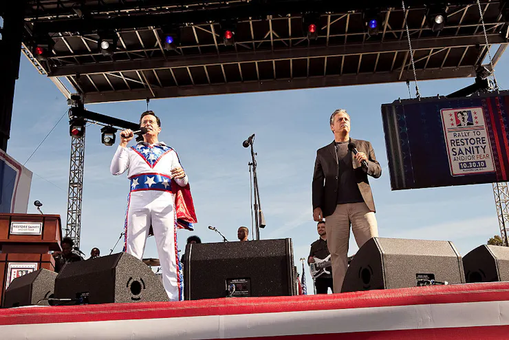 Two middle-aged men, one wearing a red, white and blue daredevil outfit with a cape, hold microphones on stage.