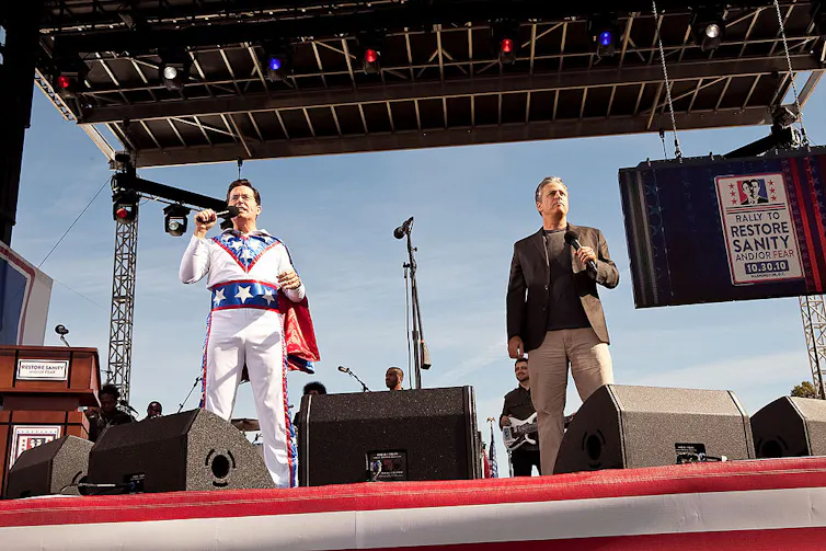 Two middle-aged men, one wearing a red, white and blue daredevil outfit with a cape, hold microphones on stage.