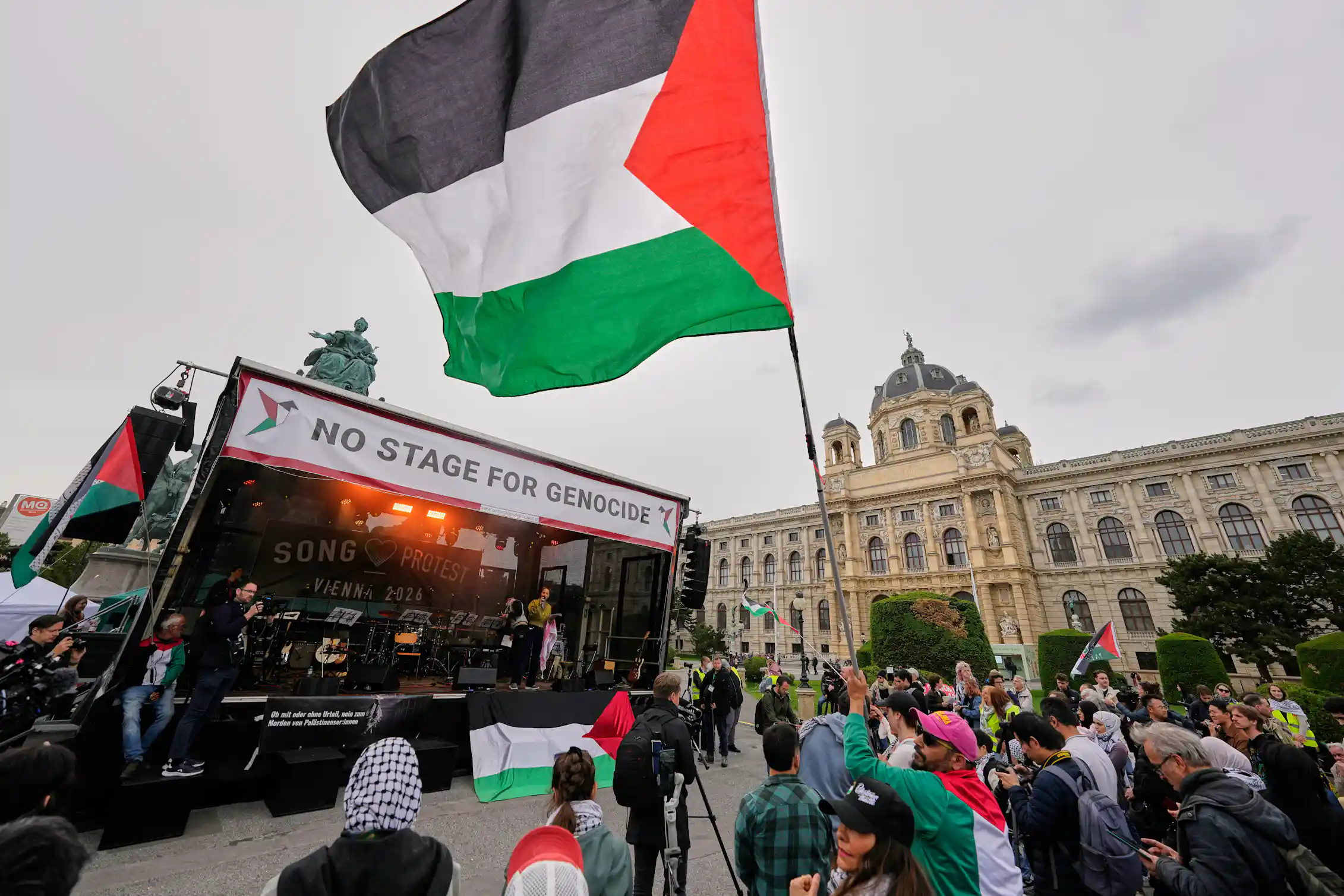 Banner above a stage reads: 'No stage for genocide'. A Palestinian flag waves above a crowd.