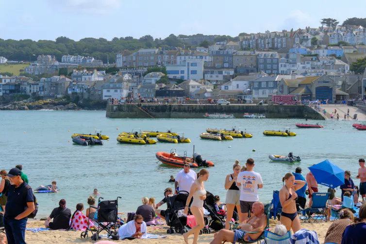 people in summer clothes and swimsuits on beach, boats and coastal village in background