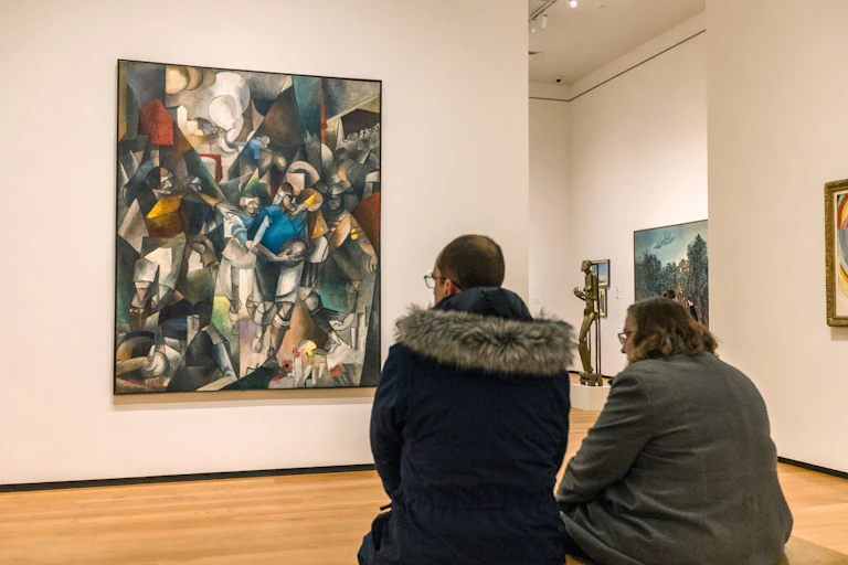 A couple sit on a bench at an art gallery looking at a painting.