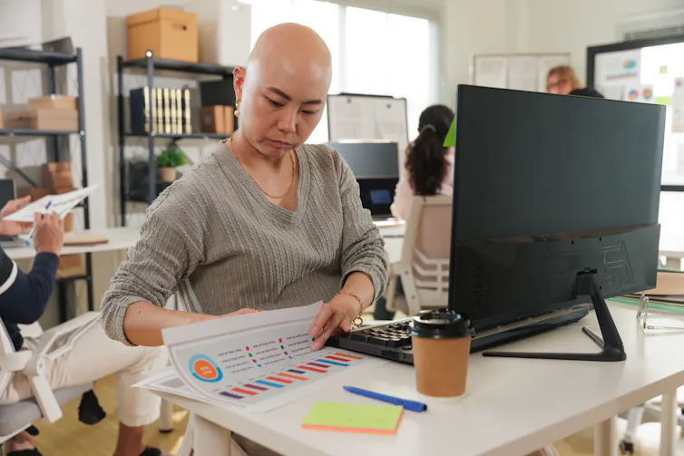 A bald Asian businesswoman sits at her desk in a bustling modern office.