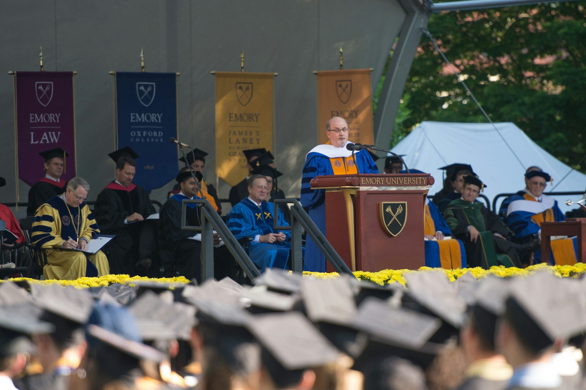 A man stands at a wooden podium on a stage, surrounded by people in graduation attire and a crowd of people wearing black graduation caps.
