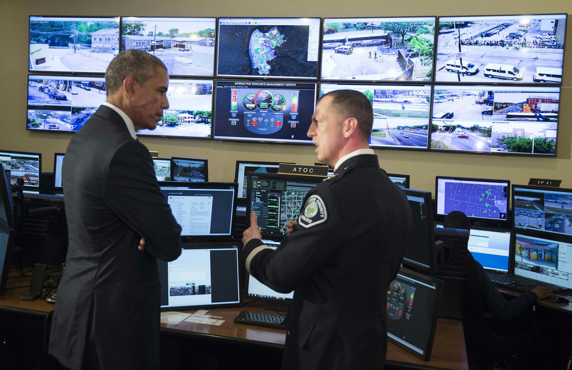 Tall man in suit talks with man in uniform as they stay in front of dozen of monitors